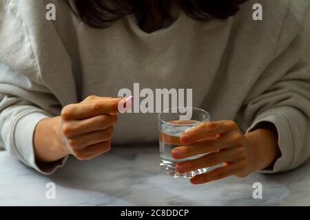 Eine Frau wird gesehen, wie sie eine Pille in der einen Hand und ein Glas Wasser auf der anderen Hand hält. Sie ist dabei, die Medikamente mit einem Schluck Wasser einzunehmen. Stockfoto