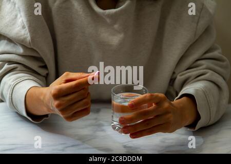 Eine Frau wird gesehen, wie sie eine Pille in der einen Hand und ein Glas Wasser auf der anderen Hand hält. Sie ist dabei, die Medikamente mit einem Schluck Wasser einzunehmen. Stockfoto