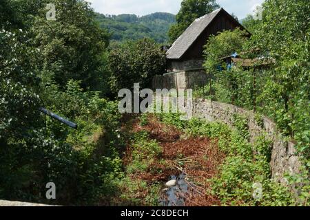 Ein altes verlassenes Dorfhaus, das fast unter wild wachsender Vegetation verloren ging. Eine Steinmauer oder Barriere schützt sie vor Strom in einem Dorf. Stockfoto