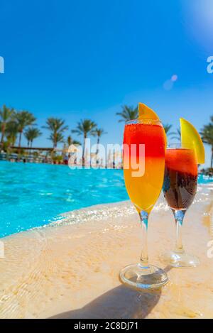 Zwei Cocktails mit natürlichem Saft und Eis am Rande des Swimmingpools. Orange Cocktail mit einer Orangenscheibe und rot mit einer Melonenscheibe mit Backdr Stockfoto