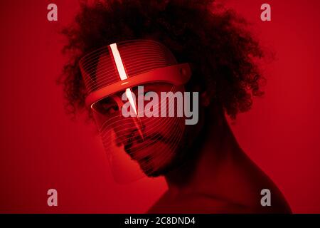 Nahaufnahme eines jungen Mannes, der eine rote LED-Lichtmaske trägt Wand zu Hause Stockfoto