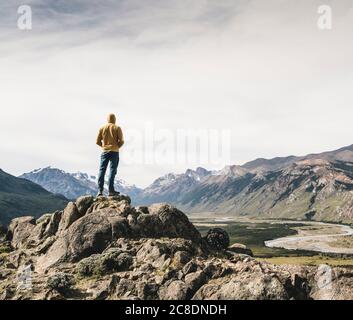 Reifer Mann mit Kapuze und Blick auf Berge gegen den Himmel, während er auf Felsen steht, Patagonien, Argentinien Stockfoto