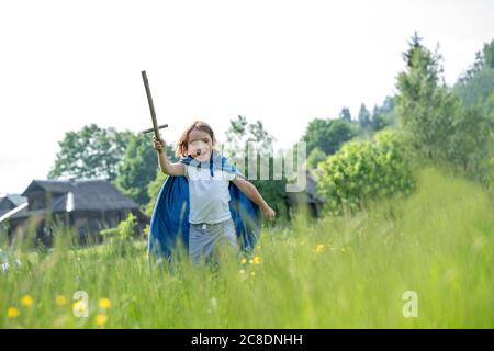 Verspielter Junge trägt Umhang mit Spielzeug Schwert beim Laufen auf Grasbewachsenes Land gegen klaren Himmel Stockfoto