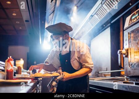 Koch trägt eine schützende Gesichtsmaske und bereitet ein Gericht im Restaurant zu Küche Stockfoto