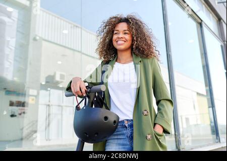 Nachdenkliche junge Frau lächelt, während mit Elektro-Push-Roller stehen In der Stadt Stockfoto