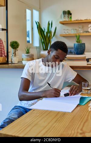 Junger Mann, der im Sitzen Notizen im Buch auf den Tisch schreibt Zu Hause Stockfoto