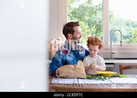 Lächelnder Mann, der beim Sitzen auf den Sohn schaut, der grüne Erbse hält In der Küche Stockfoto