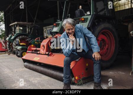 Lächelnder älterer Mann auf einem Bauernhof, der auf einem Traktor sitzt Im Stall Stockfoto