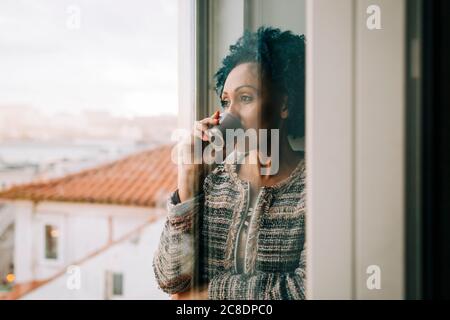 Frau, die Kaffee trinkt, während sie zu Hause durch das Fenster schaut Durch Glastür Stockfoto