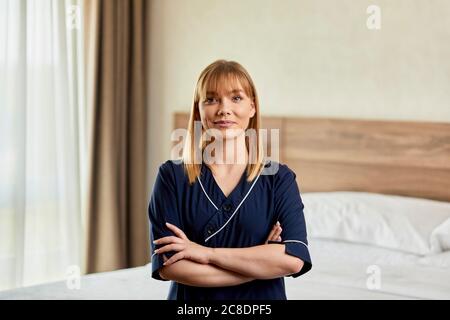 Selbstbewusstes Zimmermädchen mit gekreuzten Armen, das im Schlafzimmer des Hotels steht Stockfoto