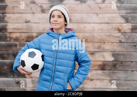 Nachdenklicher Junge in warmer Kleidung, der im Stehen den Fußball hält Gegen die Wand Stockfoto