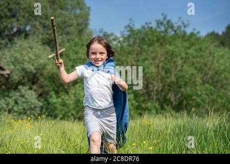 Verspielter Junge trägt Umhang mit Spielzeug Schwert beim Laufen auf Grasland Stockfoto