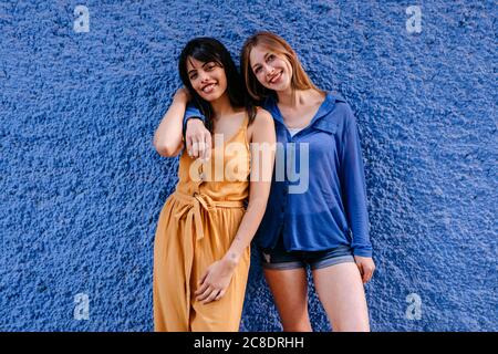 Lächelnde Freundinnen, die in der Stadt gegen die blaue Mauer stehen Stockfoto