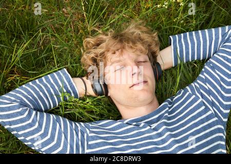 Porträt eines jungen Mannes mit lockigen blonden Haaren liegend Eine Wiese, die Musik mit Kopfhörern hört