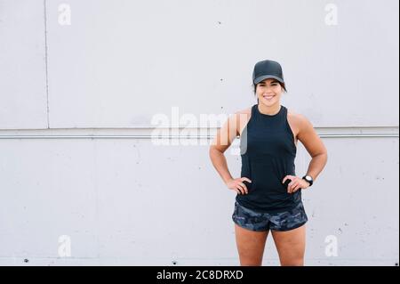 Lächelnde junge Frau mit Armen Akimbo gegen Wand in Stadt Stockfoto