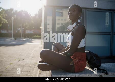 Junge Frau schaut weg, während sie auf der Stützmauer sitzt Stadt an sonnigen Tagen Stockfoto