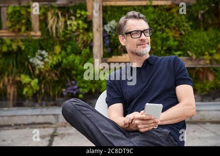 Nachdenklich reifen Mann hält Smartphone, während auf einem Stuhl sitzen Am Hof Stockfoto