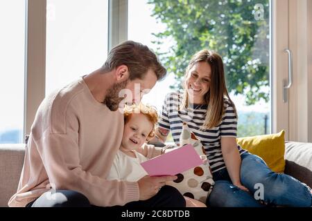 Lächelnde Frau, die sitzt und den Mann anschaut, der Bilderbuch liest Zu Jungen im Wohnzimmer Stockfoto