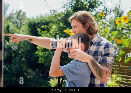 Vater zeigt dem Sohn etwas, der durch ein Fernglas auf den Hof schaut An sonnigen Tagen Stockfoto