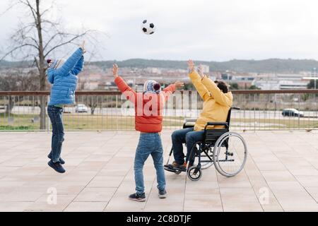 Spielerischer Vater sitzt auf dem Rollstuhl und wirft Ball auf begeisterte Söhne Im Park Stockfoto