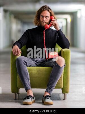 Seriöser junger Mann, der über das Telefon spricht, während er auf dem Sessel sitzt Zu Hause Stockfoto