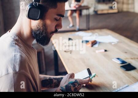Kreativer junger Geschäftsmann mit Kopfhörern und Smartphone im Loft-Büro Stockfoto