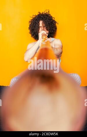 Verrückter Mann mit lockigen Haaren, der Didgeridoo spielt, während er dagegen sitzt Gelber Hintergrund Stockfoto