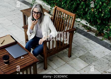Lächelnde Geschäftsfrau, die im Sitzen über einen Smartphone-Lautsprecher spricht Im Garten Stockfoto