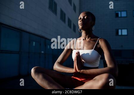 Junge Frau mit geschlossenen Augen meditiert, während sie gegen Gebäude sitzt In der Stadt Stockfoto