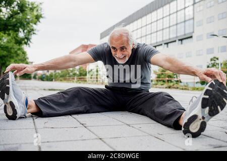 Lächelnder älterer Mann, der Körper streckt, während er auf dem Fußweg sitzt Stadt Stockfoto