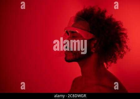 Junger Mann mit geschlossenen Augen trägt rote LED-Lichtmaske Gegen die Wand zu Hause Stockfoto