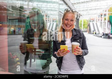 Lächelnde Frau hält Kaffeetasse mit Smartphone im Stehen Auf dem Bürgersteig in der Stadt Stockfoto
