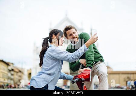 Frau, die Selfie mit ihrem Freund macht, während sie auf der Vespa in Florenz, Italien, sitzt Stockfoto