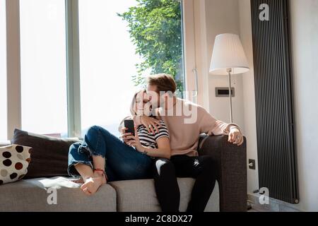 Mann küsst Frau, während er sich auf dem Sofa gegen das Fenster entspannt Wohnzimmer zu Hause Stockfoto