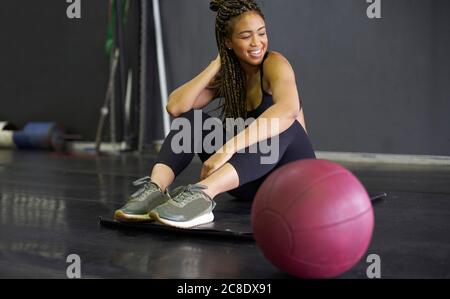 Lächelnde junge Frau mit geflochtenen Haaren sitzt auf Matte in Fitnessraum Stockfoto