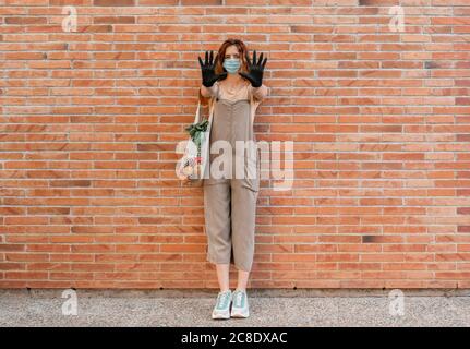 Frau trägt Maske und Handschuhe, die Hände zeigen, während sie auf steht Fußweg gegen Backsteinmauer Stockfoto