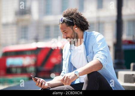 Porträt eines lächelnden jungen Mannes, der im Freien sitzt und Musik mit Handy und Kopfhörer hört, London, Großbritannien