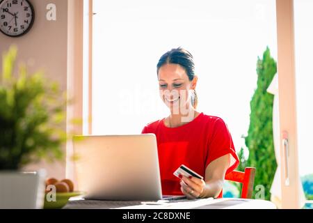 Lächelnd schöne Frau hält Kreditkarte, während mit Laptop an Esstisch am Fenster Stockfoto