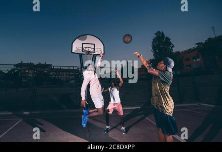 Männliche Freunde spielen Basketball auf dem Platz gegen klaren Himmel an Nacht Stockfoto