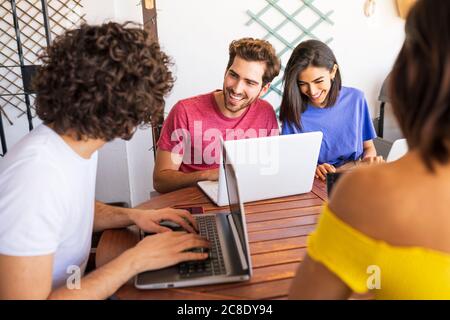 Lächelnde junge Freunde, die Laptops benutzen, während sie am Tisch sitzen Hinterhof Stockfoto