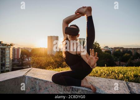 Flexible junge Frau mit VR-Brille während der Durchführung Yoga auf Stützmauer in der Stadt während des Sonnenuntergangs Stockfoto