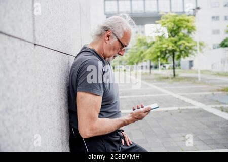 Älterer Mann, der im Stehen eine Brille mit einem Smartphone trägt Mauer in der Stadt Stockfoto
