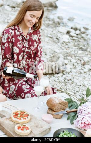 Lächelnde junge Frau, die Wein in Glas gießt, während sie ansitzt Seeufer Stockfoto