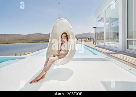 Glückliche Frau, die in einem Hängesessel über dem Pool saß und sprach Am Telefon Stockfoto