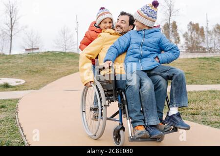 Glücklicher Mann mit verspielten Söhnen, die auf dem Fußweg im Rollstuhl reiten parken Stockfoto