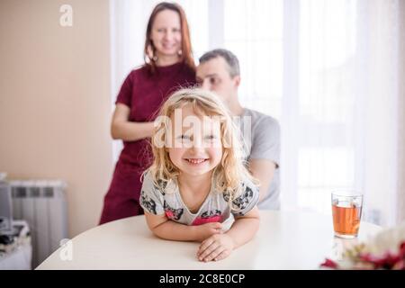 Happy nettes Mädchen lehnte sich auf Esstisch mit Eltern stehen Im Hintergrund zu Hause Stockfoto