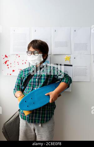 Junge trägt Maske hält Skateboard, während sie gegen Wand in Schule Stockfoto