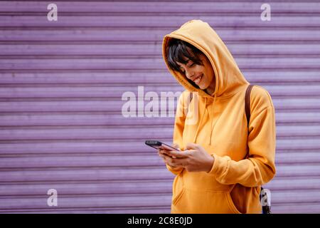 Portrait einer jungen Frau mit gelbem Hoodie beim Smartphone-Check in Vorderseite des violetten Hintergrundes Stockfoto