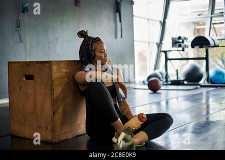 Nachdenkliche Sportlerin, die Musik hört, während sie auf dem Boden sitzt Fitnessraum Stockfoto