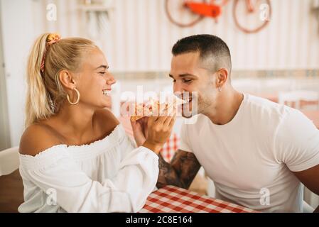 Fröhliche junge Frau Fütterung Pizza zu Freund, während im Sitzen Restaurant Stockfoto